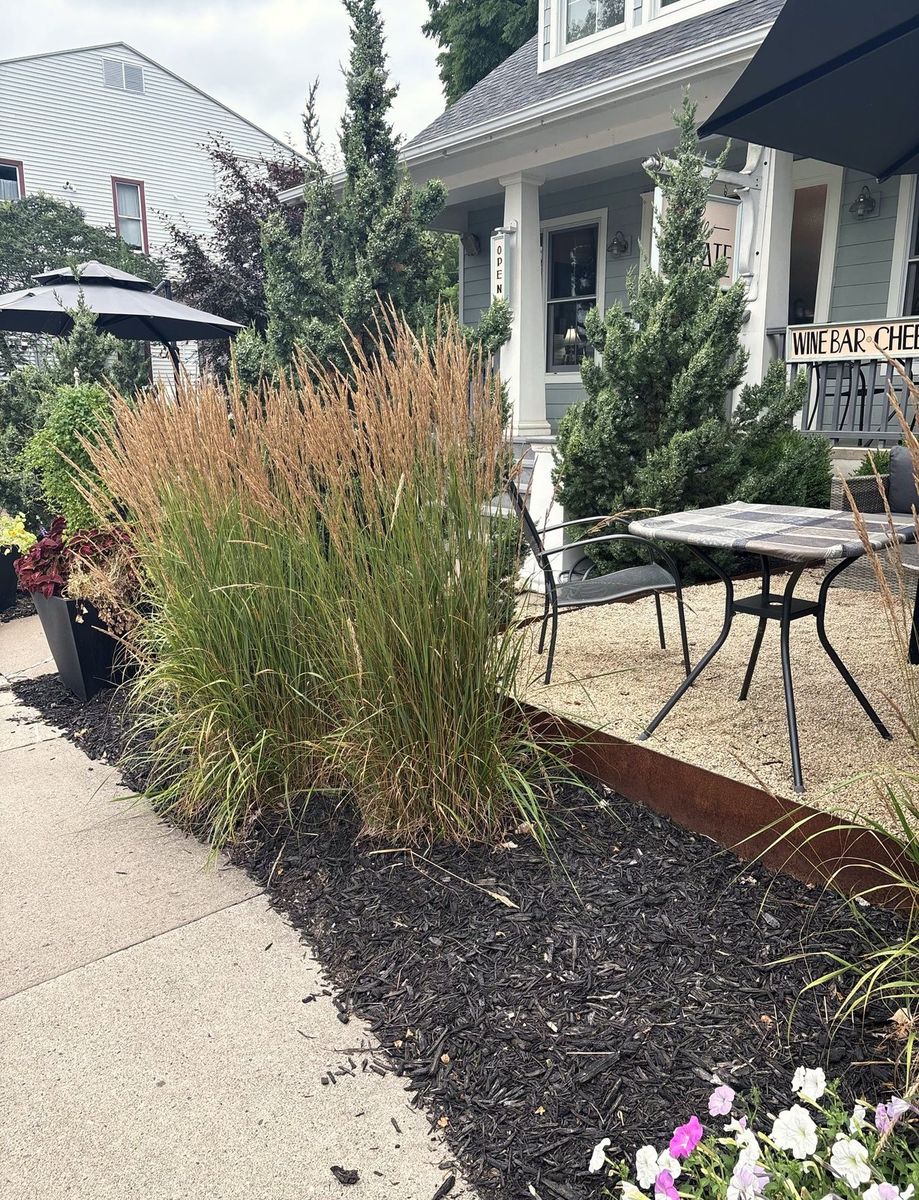 Ornamental grasses and dark mulch bordering a gravel patio with cafe tables outside a shop.