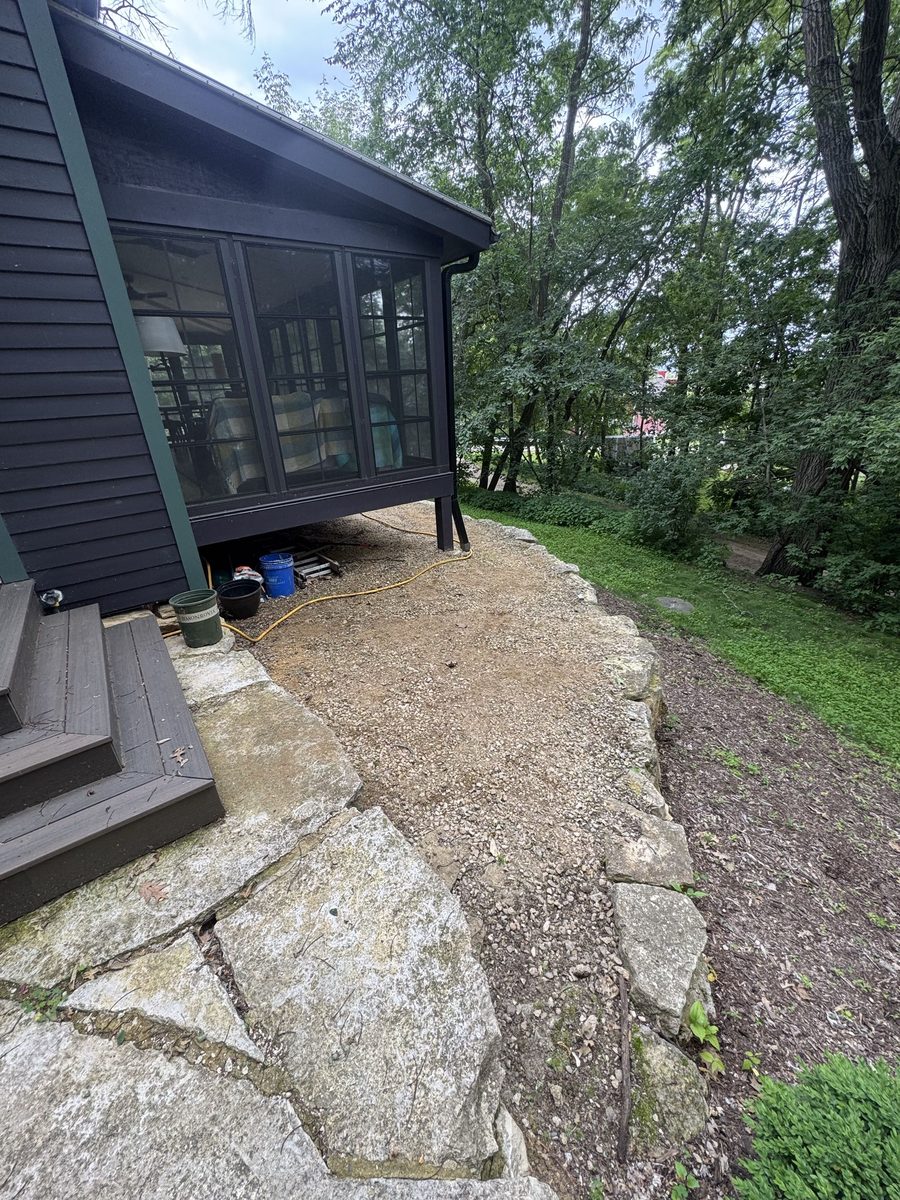 A gravel area beside a screened porch, edged with boulders and mulch, with flagstone at one side and woods beyond.