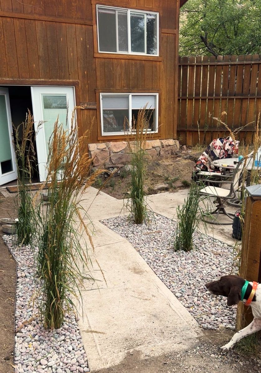 A concrete walkway leading to a back door, edged with river rock beds and tall ornamental grasses.
